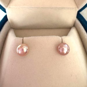 May include: A pair of pink pearl stud earrings displayed inside a light beige jewelry box. The pearls have a soft, iridescent sheen and are round in shape. The jewelry box has a teal blue velvet exterior.