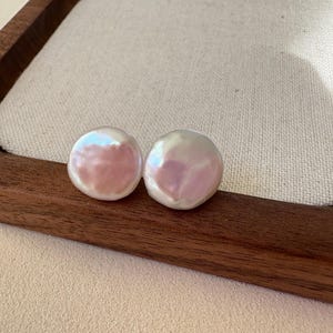 May include: Two round, iridescent pearl earrings with a pink and white hue. The earrings are displayed on a wooden tray with a beige fabric insert. The pearls have a smooth, polished surface.