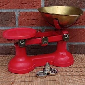 Peut inclure: Une balance rouge vintage avec un bol en laiton et une petite plateforme. La balance comprend un ensemble de poids. La balance est posée contre un mur de briques.
