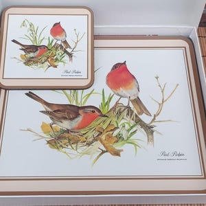May include: A set of decorative placemats and a coaster featuring illustrations of red robins perched on branches with greenery. The placemats and coaster have a cream background with a brown border and are presented in a white box.