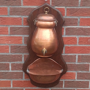 Könnte beinhalten: Ein wandmontierter Wasserspender aus Kupfer mit gehämmertem Finish, einem Deckel mit Messingringgriff und einem kleinen Zapfhahn. Unter dem Spender befindet sich ein passendes Kupferbecken, beides auf einer dunkelbraunen Holzplatte montiert.