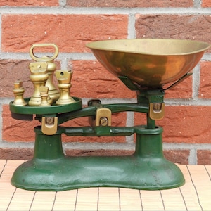 Peut inclure: Une balance de style vintage dotée d'une base et d'un cadre en fonte vert foncé. La balance comprend un grand bol en laiton poli sur le côté droit et une plate-forme circulaire sur la gauche contenant un ensemble de cinq poids en laiton gradués.