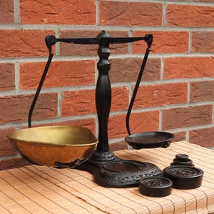 May include: A black cast iron balance scale featuring a central vertical post, a horizontal beam, and two hanging pans. One pan is made of polished brass, while the other is black metal. Three circular black iron weights are placed near the base.
