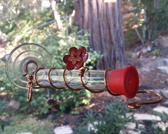 May include: A glass hummingbird feeder with a red flower design and a red plastic cap. The feeder is attached to a suction cup and has a copper wire coil.