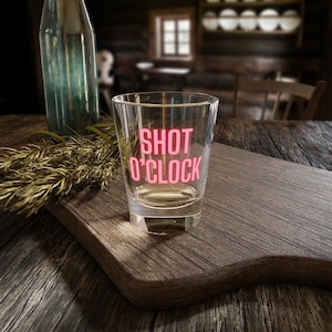 May include: A clear shot glass with the words "SHOT O'CLOCK" in pink lettering. The glass is on a wooden cutting board with a bottle and dried wheat in the background. The shot glass is empty.