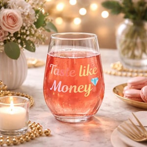 Puede incluir: Una copa de vino sin pie llena de una bebida rosa. La copa tiene la inscripción "Taste like Money" en letras coloridas, con un diamante azul. La copa está sobre una mesa con flores, una vela y cuentas doradas.