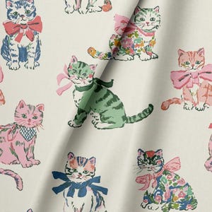 May include: A light-colored fabric featuring a repeating pattern of illustrated cats in various colors, including pink, green, and blue. Each cat is adorned with a bow or a patterned neck accessory, rendered in a hand-drawn style with visible ink lines.