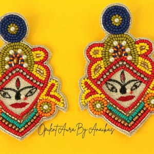 May include: A pair of beaded earrings with a face design, featuring red, yellow, orange, and blue beads. The earrings have a silver outline and a blue circular top. The background is a solid yellow.