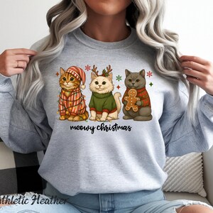 May include: Athletic Heather sweatshirt featuring three cartoon cats dressed for Christmas. One cat wears a striped hat and lights, another a green sweater, and the third a red plaid sweater and holds a gingerbread man. The text "meowy christmas" is below.