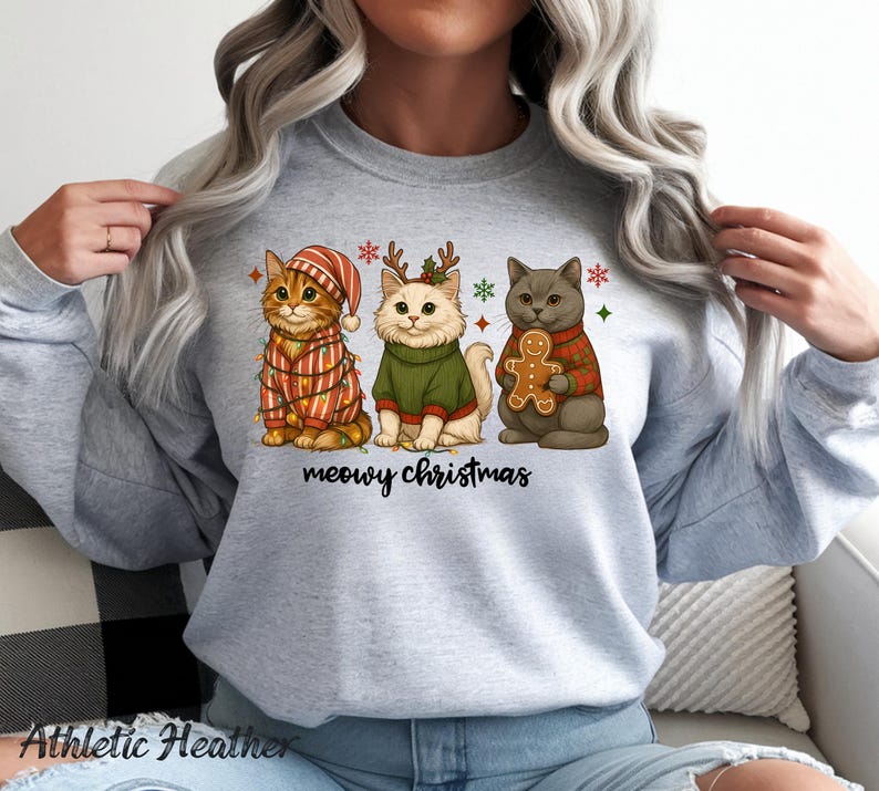 May include: Athletic Heather sweatshirt featuring three cartoon cats dressed for Christmas. One cat wears a striped hat and lights, another a green sweater, and the third a red plaid sweater and holds a gingerbread man. The text "meowy christmas" is below.