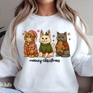 May include: White sweatshirt featuring a festive design with three cartoon cats. The cats are dressed in holiday attire, including a striped hat, a green sweater, and a plaid sweater. The text "meowy christmas" is written in black script below the cats.