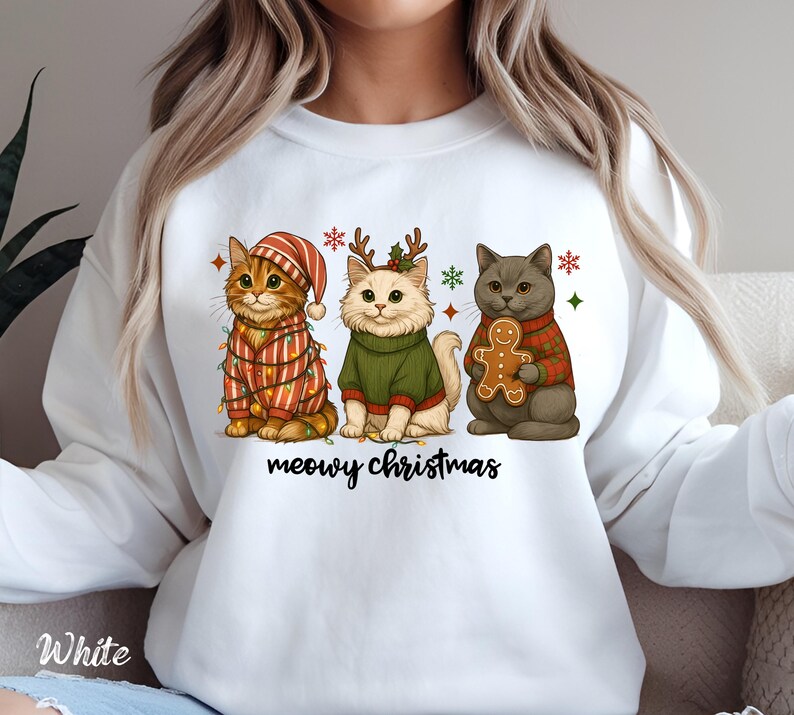 May include: White sweatshirt featuring a festive design with three cartoon cats. The cats are dressed in holiday attire, including a striped hat, a green sweater, and a plaid sweater. The text "meowy christmas" is written in black script below the cats.