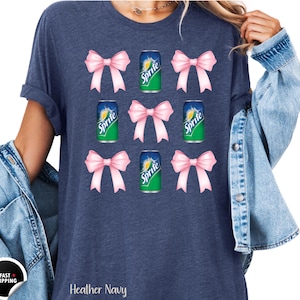 May include: Heather navy blue t-shirt with a design featuring pink bows and Sprite soda cans. The text "Sprite" is visible on the cans. The shirt is made of a soft material.