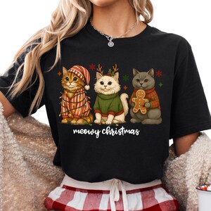 May include: Black t-shirt featuring three cartoon cats dressed for Christmas. The cats wear festive sweaters, hats, and hold a gingerbread man. The text "meowy christmas" is written below the cats.