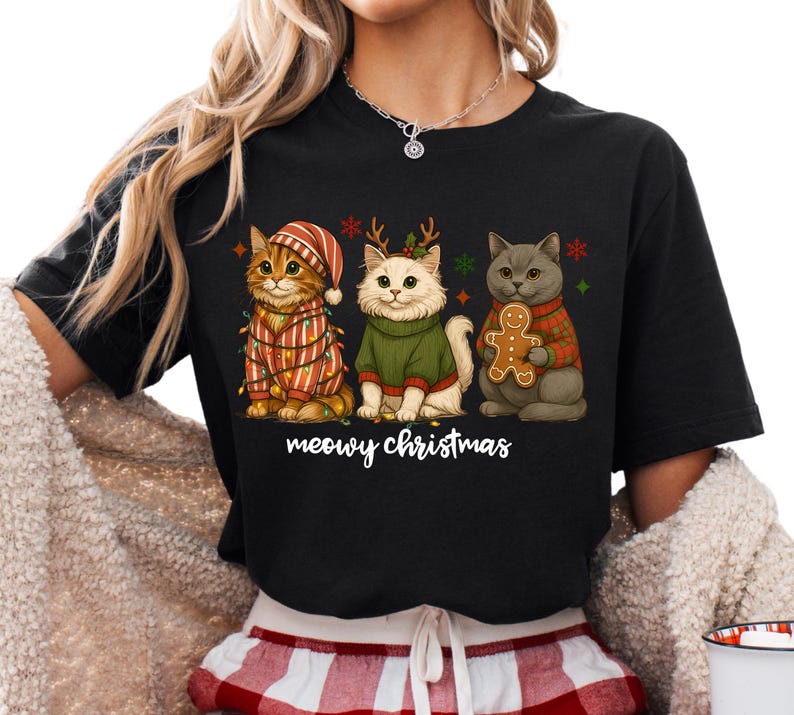 May include: Black t-shirt featuring three cartoon cats dressed for Christmas. The cats wear festive sweaters, hats, and hold a gingerbread man. The text "meowy christmas" is written below the cats.