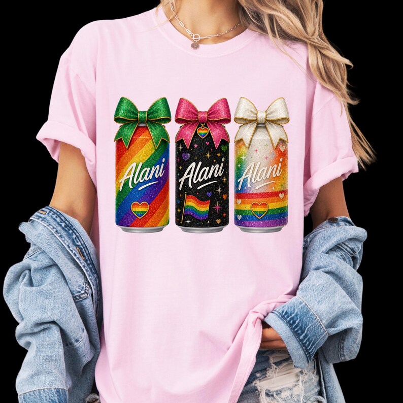 Pu&ograve; includere: T-shirt rosa con tre lattine colorate con fiocchi. Le lattine hanno sfondi arcobaleno, neri e bianchi con la scritta "Alani" e un design arcobaleno a forma di cuore. La maglietta &egrave; indossata da una persona.