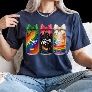 Pu&ograve; includere: T-shirt blu navy con tre lattine colorate. Ogni lattina presenta un design arcobaleno, la parola "Alani" e un cuore. Le lattine hanno fiocchi verdi, rosa e crema. La lattina centrale ha uno sfondo nero con stelle e una bandiera arcobaleno.