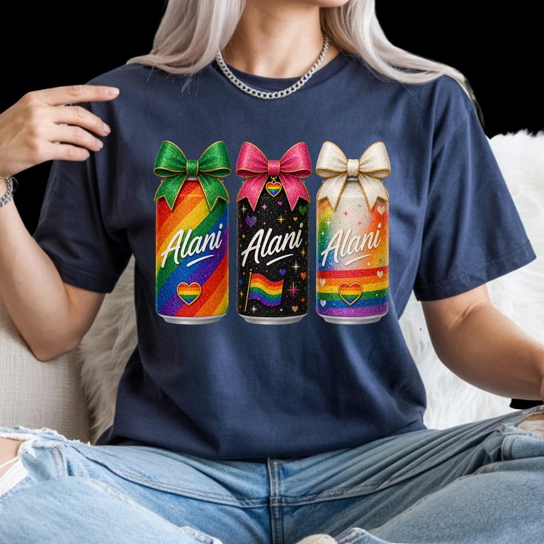 Pu&ograve; includere: T-shirt blu navy con tre lattine colorate. Ogni lattina presenta un design arcobaleno, la parola "Alani" e un cuore. Le lattine hanno fiocchi verdi, rosa e crema. La lattina centrale ha uno sfondo nero con stelle e una bandiera arcobaleno.