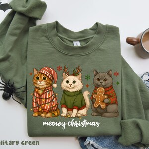 May include: Military green sweatshirt featuring three cartoon cats in festive attire. One cat is wrapped in Christmas lights, another wears a sweater and antlers, and the third holds a gingerbread man. The text "meowy christmas" is printed below.