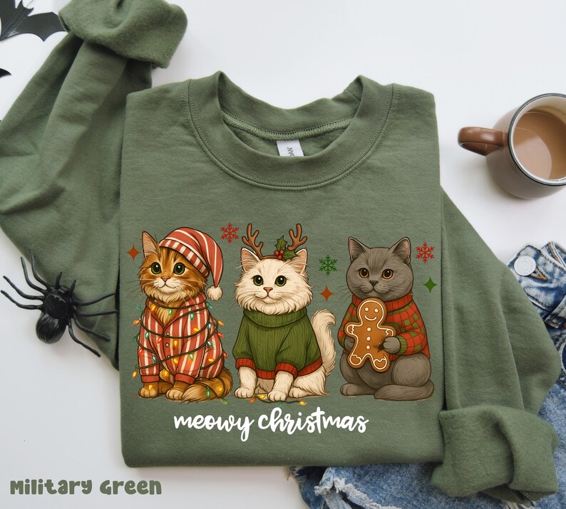May include: Military green sweatshirt featuring three cartoon cats in festive attire. One cat is wrapped in Christmas lights, another wears a sweater and antlers, and the third holds a gingerbread man. The text "meowy christmas" is printed below.