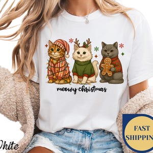 May include: White t-shirt featuring a festive design with three cartoon cats. The cats are dressed in holiday attire, including sweaters and a hat, with Christmas lights and snowflakes. The text "meowy christmas" is below the cats.