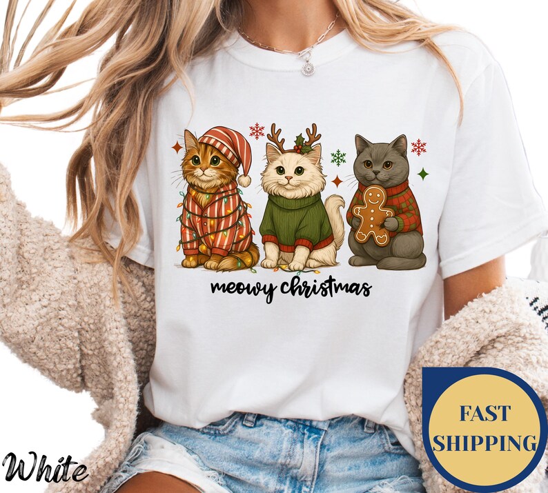 May include: White t-shirt featuring a festive design with three cartoon cats. The cats are dressed in holiday attire, including sweaters and a hat, with Christmas lights and snowflakes. The text "meowy christmas" is below the cats.