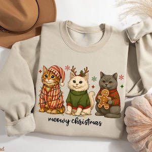 May include: Beige sweatshirt featuring three cartoon cats dressed for Christmas. One cat wears a striped hat and is wrapped in lights. Another wears a green sweater and reindeer antlers. The third cat holds a gingerbread man. The text "meowy christmas" is below.