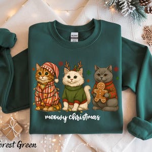 May include: Forest green sweatshirt featuring three cartoon cats dressed for Christmas. One cat wears a striped hat and is wrapped in lights. Another wears a green sweater and antlers. The third cat holds a gingerbread man. The text "meowy christmas" is below.