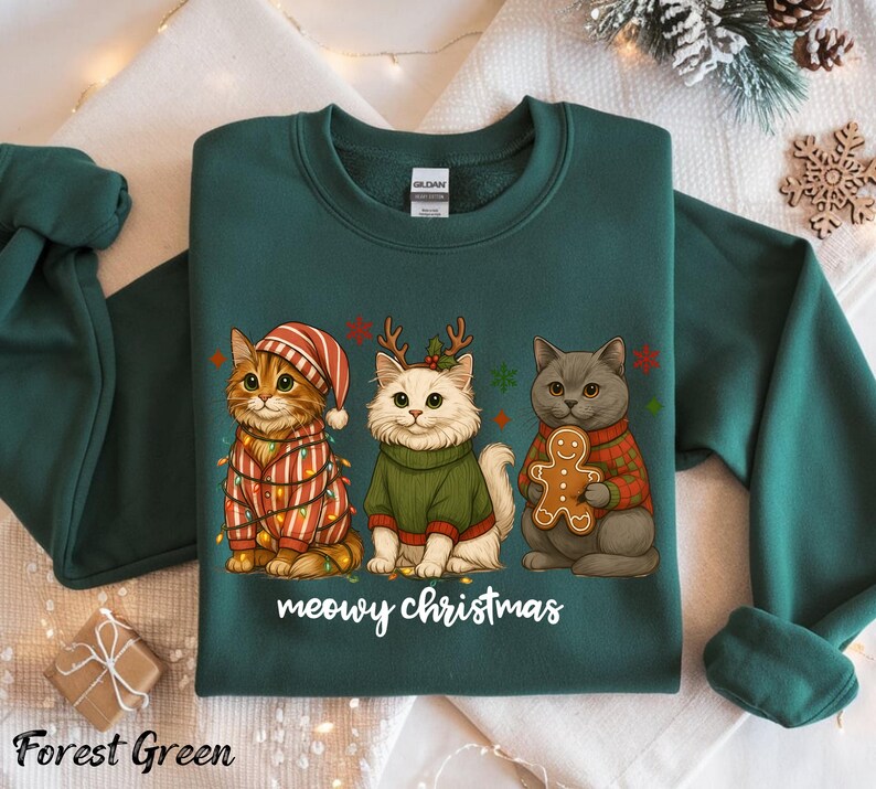 May include: Forest green sweatshirt featuring three cartoon cats dressed for Christmas. One cat wears a striped hat and is wrapped in lights. Another wears a green sweater and antlers. The third cat holds a gingerbread man. The text "meowy christmas" is below.