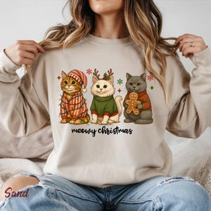May include: Beige sweatshirt featuring three cartoon cats dressed for Christmas. One cat wears a striped hat and sweater with lights, another a green sweater and antlers, and the last a red plaid sweater holding a gingerbread man. The text "meowy christmas" is below.