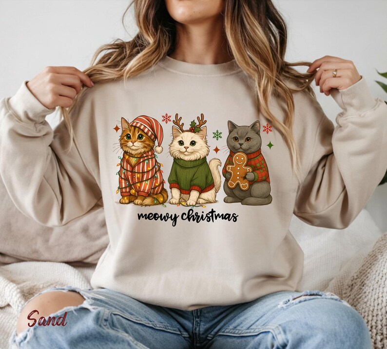 May include: Beige sweatshirt featuring three cartoon cats dressed for Christmas. One cat wears a striped hat and sweater with lights, another a green sweater and antlers, and the last a red plaid sweater holding a gingerbread man. The text "meowy christmas" is below.