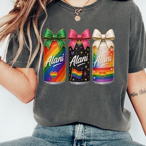 Pu&ograve; includere: Una maglietta grigia con tre lattine colorate con fiocchi. Le lattine hanno disegni arcobaleno, neri e bianchi, con la parola "Alani" in corsivo. Ogni lattina ha un cuore arcobaleno e una bandiera arcobaleno.