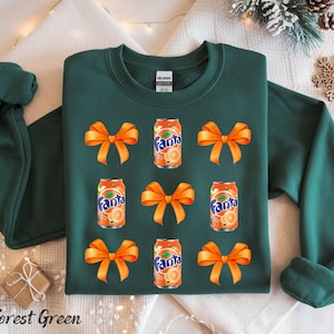 May include: Forest green sweatshirt with a festive design. The sweatshirt features orange bows and Fanta soda cans. The text "Fanta" is visible on the cans. The sweatshirt is a comfortable, casual garment.