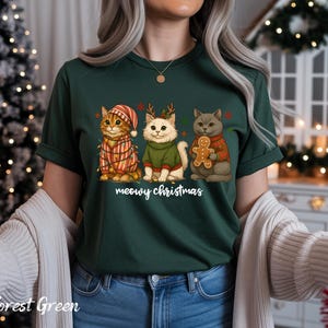 May include: Forest green t-shirt featuring three cartoon cats dressed for Christmas. One cat wears a striped hat and lights, another has antlers and a green sweater, and the third holds a gingerbread man. The text "meowy christmas" is below the cats.