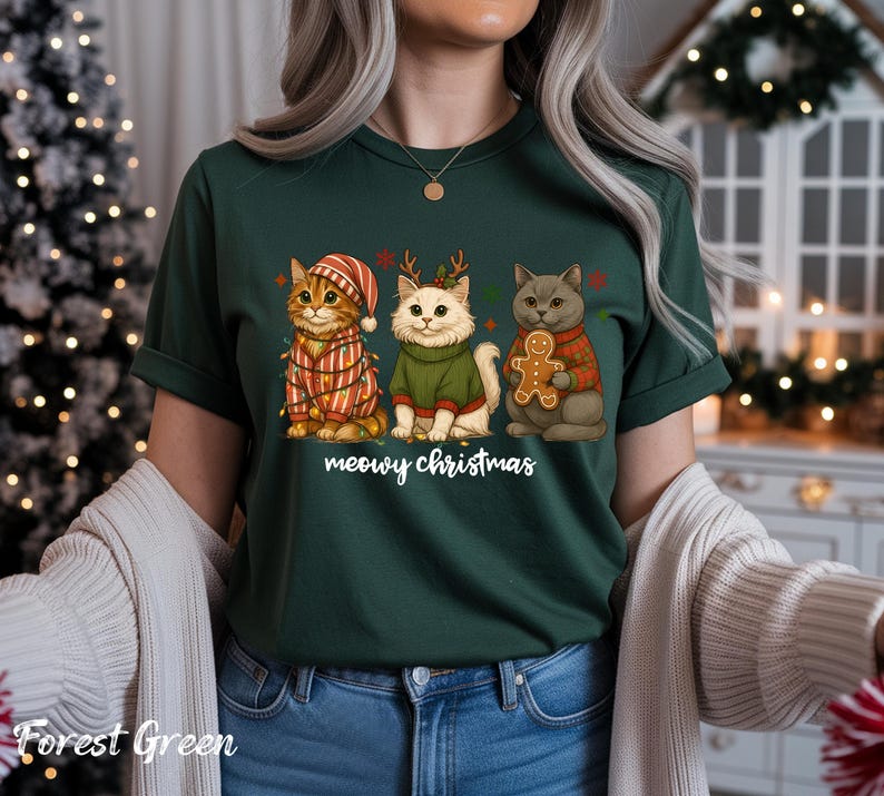 May include: Forest green t-shirt featuring three cartoon cats dressed for Christmas. One cat wears a striped hat and lights, another has antlers and a green sweater, and the third holds a gingerbread man. The text "meowy christmas" is below the cats.