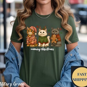 May include: Military green t-shirt featuring three cartoon cats dressed for Christmas. One cat wears a Santa hat and a striped sweater, another wears a green sweater and reindeer antlers, and the third holds a gingerbread man. The text "meowy christmas" is below.