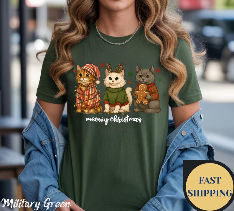 May include: Military green t-shirt featuring three cartoon cats dressed for Christmas. One cat wears a Santa hat and a striped sweater, another wears a green sweater and reindeer antlers, and the third holds a gingerbread man. The text "meowy christmas" is below.