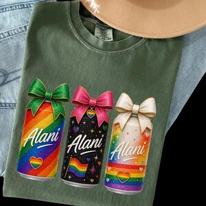 Pu&ograve; includere: T-shirt verde con tre lattine colorate con fiocchi. Ogni lattina ha un design arcobaleno, il nome "Alani" e un cuore arcobaleno. Una lattina &egrave; nera con una bandiera arcobaleno. Un cappello beige e jeans blu sono sullo sfondo.