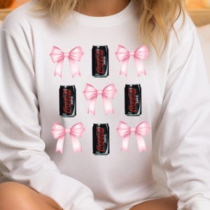 May include: White long-sleeve sweatshirt featuring a pattern of pink bows and black Coca-Cola Zero cans. The Coca-Cola cans are black with white text. The bows are a soft pink color.