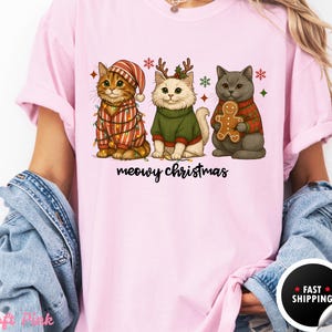 May include: Pink t-shirt featuring three cartoon cats dressed for Christmas. One cat wears a striped outfit with lights, another a green sweater and antlers, and the third a red sweater holding a gingerbread man. The text "meowy christmas" is below.