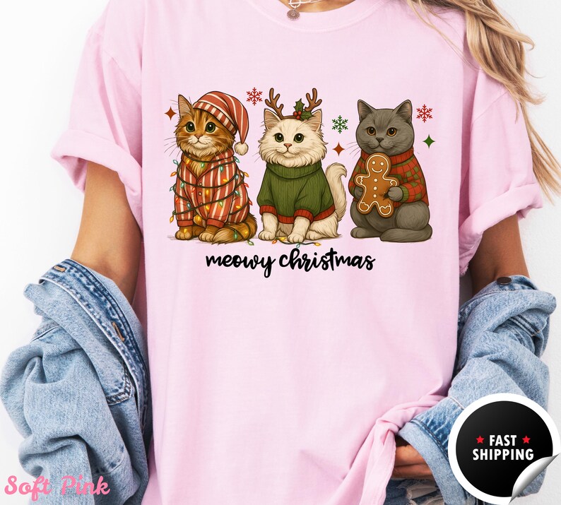 May include: Pink t-shirt featuring three cartoon cats dressed for Christmas. One cat wears a striped outfit with lights, another a green sweater and antlers, and the third a red sweater holding a gingerbread man. The text "meowy christmas" is below.