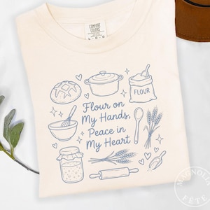 May include: Off-white t-shirt with a blue illustration of baking-related items. The text "Flour on My Hands, Peace in My Heart" is displayed. Includes a loaf of bread, pot, flour sack, and rolling pin.