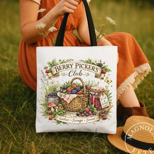 May include: White tote bag with black handles featuring a "Berry Pickers Club" design. The design includes a basket of berries, a jar of jam, and the words "Gather, Forage, Savor". The bag is held by a person sitting in a grassy field.