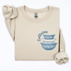 May include: A beige sweatshirt featuring a stack of three blue and white floral patterned bowls. The word "collector" is written in a cursive font above the bowls. The sweatshirt has a crew neck and long sleeves.