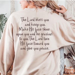 Könnte beinhalten: Beiges Sweatshirt mit dem Text: "The Lord bless you and keep you. Make His face shine upon you and be gracious to you. The Lord turn His face toward you and give you peace." Das Kleidungsstück ist neutral gefärbt.