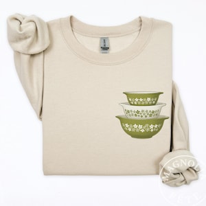 Może przedstawiać: Beżowa bluza z nadrukiem trzech zielono-białych misek do mieszania we wzory kwiatowe. Miski są w różnych rozmiarach, największa na dole i najmniejsza na górze. Bluza ma okrągły dekolt i długie rękawy.
