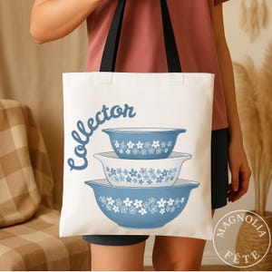 May include: White tote bag with black handles featuring a blue floral design and the word "collector" in a script font. The design includes three nested blue bowls with white floral patterns. The bag is held by a person.