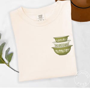 May include: Cream-colored t-shirt with a graphic of three green and white floral patterned mixing bowls. The shirt has a Comfort Colors tag and is on a white surface. A brown hat is visible in the background.