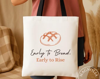 May include: A white tote bag with black shoulder straps featuring a line-art illustration of a round loaf of bread in orange. Below the bread, the text reads Early to Bread. Early to Rise in dark gray and orange lettering on the front.