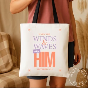 Könnte beinhalten: Weißer Tragetasche mit schwarzen Griffen. Auf der Tasche steht der Text "EVEN THE WINDS & WAVES obey HIM" in Lila und Orange, darunter "MATTHEW 8:27". Das Design enthält Sternakzente.
